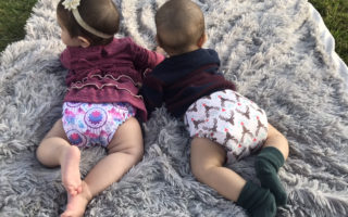 two toddlers laying on fuzzy gray blankets, facing away from camera and wearing colorful shirts with cloth diapers