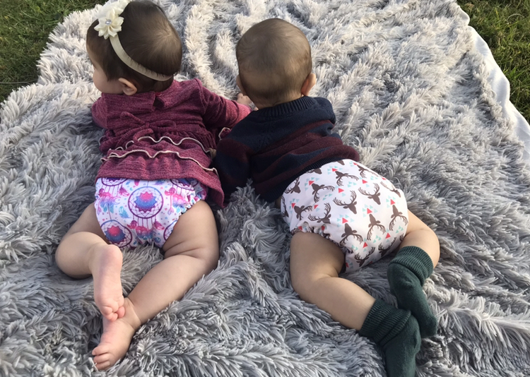 two toddlers laying on fuzzy gray blankets, facing away from camera and wearing colorful shirts with cloth diapers